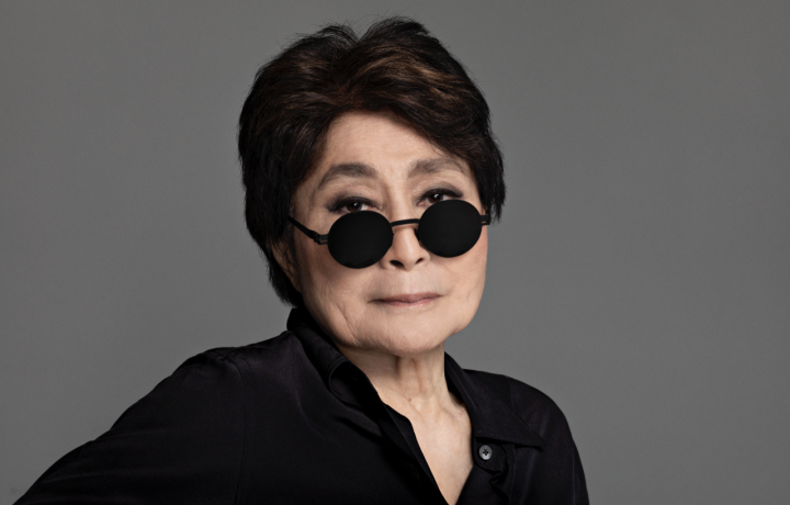 Portrait of Yoko Ono in front of a dark gray backdrop. She has black circular glasses resting on the bridge of her nose. She is wearing a black button up, has on thick eyeliner, and has a small smirk on her face. 