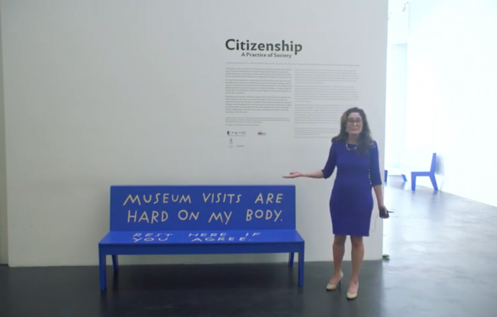 Image of Miranda Lash standing in a blue dress in a gallery next to a bright blue bench created by artist Shannon Finnegan that reads in hand painted letters: "MUSEUM VISITS ARE HARD ON MY BODY REST HERE IF YOU AGREE,"