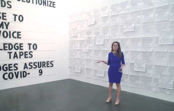 Image of Miranda Lash standing in a bright blue dress in a gallery in front of the work of Paul Ramirez Jonas. His work shows marquee letters on the left of the image, and on the right Miranda stands in front of a gallery wall with dozens of papers attached to the wall. 