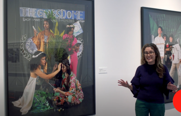 mage of Miranda Lash standing in a gallery wearing a navy blue sweater. She stands to the right of a photograph from Adelita Husni-Bey's work titled "The institution will be run by a morally superior Al."