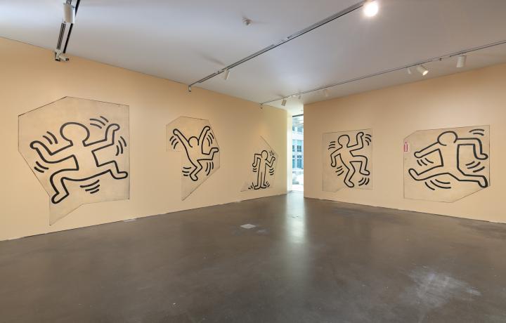 Corner of a gallery in MCA Denver featuring work by Keith Haring. The floors are concrete and the walls are a yellowish beige, and feature works that look like black outlines of figures dancing, with lines around them that suggest movement. 
