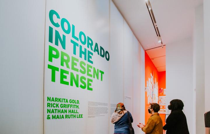 Side view of a gallery at MCA Denver with bright green wall text on white panels that reads, "Colorado in the Present Tense: Narkita Gold, Rick Griffith, Nathan Hall, & Maia Ruth Lee” with smaller, illegible black text next to it. Three people stand in front of the wall text reading, and a peek into a gallery with tomato red walls can be seen.  