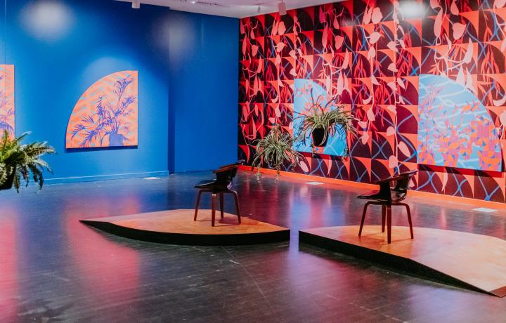 Corner of a clorful gallery space. One wall is cobalt blue, another wall features wallpaper that looks like red squares, red leaves, and thin blue netting. On the walls are works depicting silohettes of plants in similar colors and designs as featured on the wallpaper. In the center of the room are two platforms with midcentury black leather chairs on them, and there are spider plants suspended from the ceiling. 