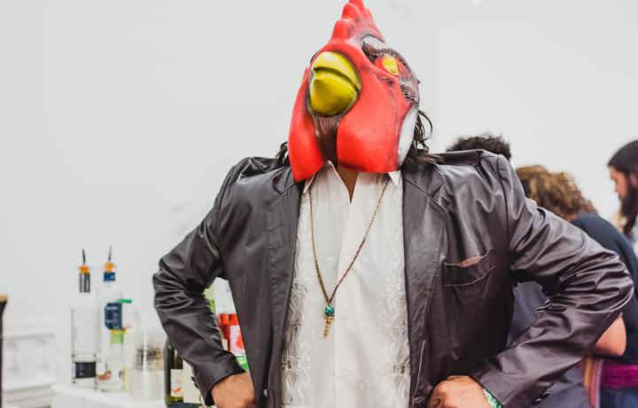 Person standing in front of a bar with their hands on their hips, wearing a bright red bird mask on their head. 