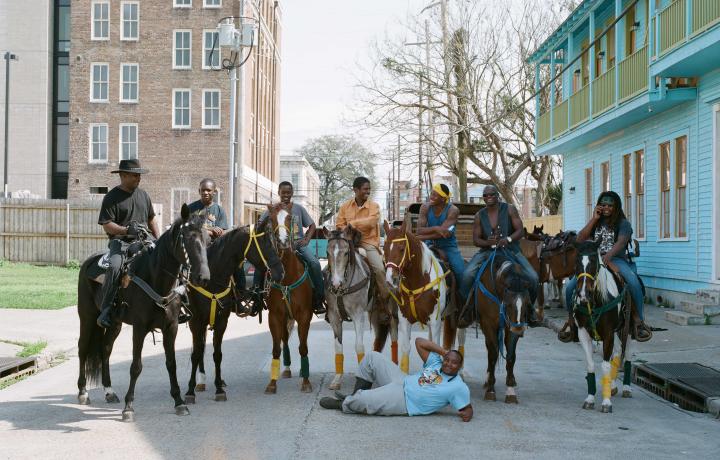  Image of seven people on horseback, all standing in the street. There is one person laying on the street in front of the horses.