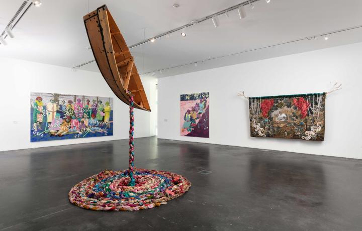 Three colorful artworks on the wall, one suspended from the ceiling in an MCA Denver gallery. The one suspended from the ceiling is a boat with vintage saris spilling out.