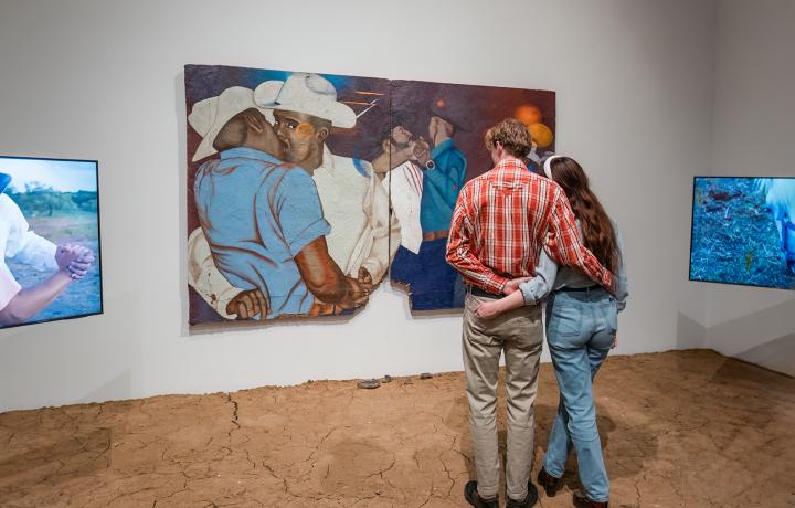 Two people with their backs towards the camera, standing with their arms around each other. They are looking at a large painting on the wall.