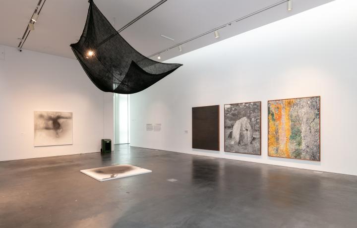 Gallery featuring a large burlap sack hanging from the ceiling. There are three large works of art hung on the wall.