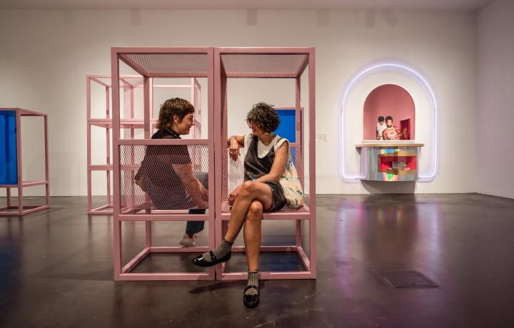 Two people sitting in a gallery on a light pink sculpture.