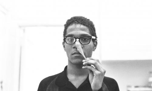 Black and white portrait of Jean Michel Basquiat. He is wearing glasses and appears to be pulling a putty off of his face