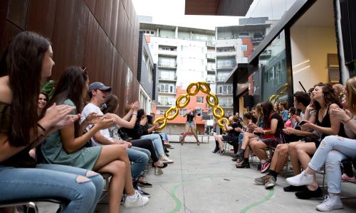 Young adults are seating in an alleyway watching a performer at the end of the alley way. There are golden balloons resembling a chain at the stage. 