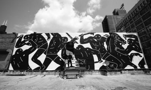 Dramatic black and white shot of a man posing in front of a mural depicting abstract figures engaging in violence. 