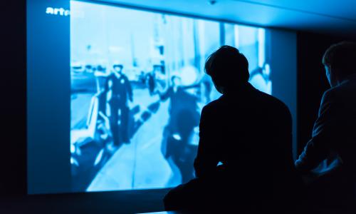 Two people are seated on a bench in a dark room. A video is projected onto a wall depicting police in an unknown outdoor environment. 