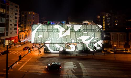 Exterior shot of the MCA Denver at night being lit by spotlights. The building is wrapped in art by Cleon Peterson depicting abstract figures fighting.