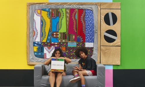 Two young adults sit on a couch in front of a colorful wall. 