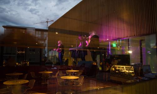 A band performs basked in pink lights through the reflection of glass. Round tables are visible through the glass