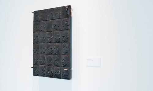 Installation shot of an artwork hanigng on a wall. it is black and made up of grids depicting portraits of people. 