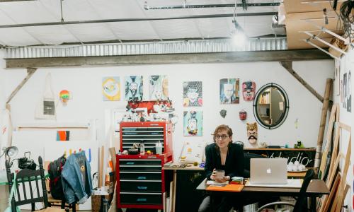 Sierra at her desk in her studio space. She is surrounded by several artworks. A mac book is open on her desk and she holds a ceramic mug in her hands. 