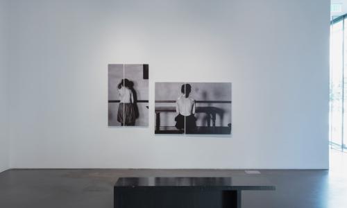 Installation shot of two photographs with a line down the middle. They depict two topless women facing away from the camera. 