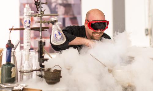 A man is wearing black safety goggles. To his side are chemical beakers. He is looking and smiling into a cloud of fog produced by dry ice. 