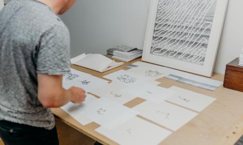 Candid shot of a man looking at several sketches that rest on a wood table. 