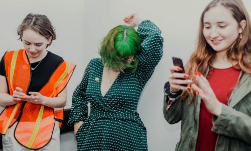A candid shot of three young women in front of a blank white background. One is wearing an orange safety vest, the other is mid dance with green hair, and the other looks at her phone, smiling. 