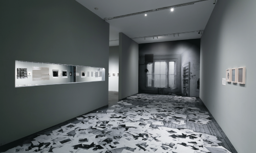 Shot of a gallery space. The floor and far wall are covered in a vinyl wrap. The floor depicts a hardwood floor covered in papers while the far wall depicts a rustic window. 