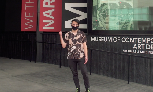 Image: Young man in a mask stands outside the museum and he stands in a printed shirt, black pants, and motions with his right hand to follow him.