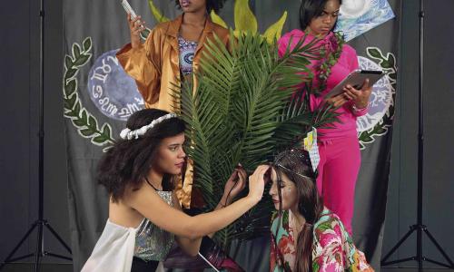 Four young women of different racial backgrounds surround a fern in an indoor space. They are dressed colorfully while donning avante garde makeup. They are posed in front of a hand painted background with imagery of plants and zodiac symbols while two of them look at iPads.  