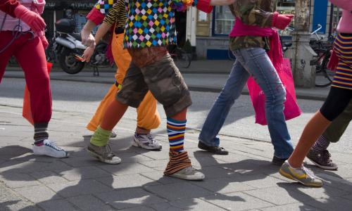 Candid photo of six people marching from the shoulders down in an outdoor urban space. They are wearing a wide variety of clothes from jeans to sweat to shorts, in clashing and bright patterns. 