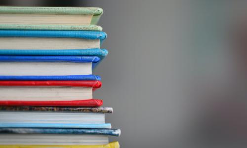A close up shot of seven colorful worn books stacked ontop of eachother. The background is blurred and indistinguishable. 
