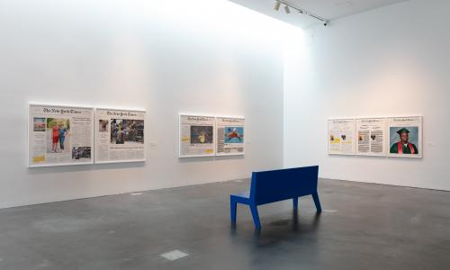 A white museum gallery with concrete floors. There are seven large, framed newspapers hung on the two walls. Near the center, there is a blue bench, and only the back of the bench is visible. 