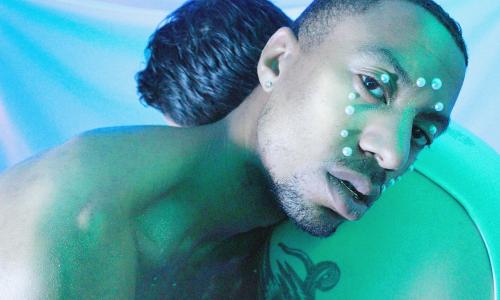 A photo of two men facing one and laying on one another’s shoulder. Only one of their faces is in view, and it’s covered in pearls. The photo looks as if it was taken underneath a blue/green hue and the backdrop is comprised of the colors blue, purple, and green.