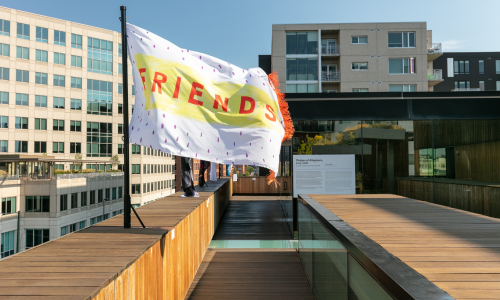 A flag flying on the MCA Denver rooftop, next to a row of other flags fixed to the light brown wood ledge around the perimeter of the rooftop. The flag in view has the word “FRIENDS” on it in bold, red text, and is printed on a yellow block of color. Behind the yellow block of color are purple polka dots on a white background, with red fringe decorating the right side of the flag.