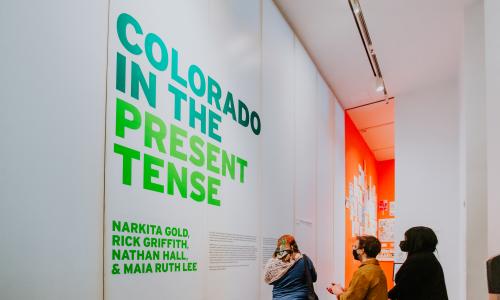 Image of a gallery with bright green wall text that reads Colorado in the Present Tense. Three people stand in front of the wall text reading.