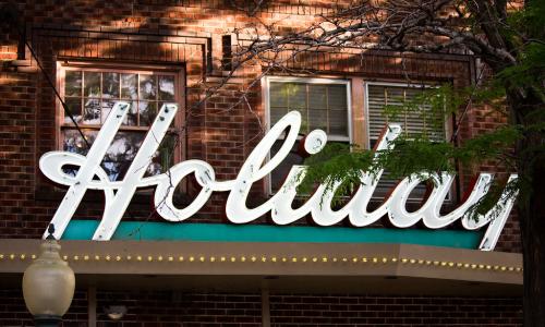 Close up street view of a sign on a brick building that reads “Holiday” in white, cursive lettering.  Behind the sign are three windows, and below are marquee lights. 
