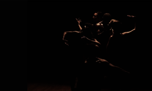 dark image of performers in pose. Lighting is rimming their arms, distinguishing them from the dark shadows of the photo