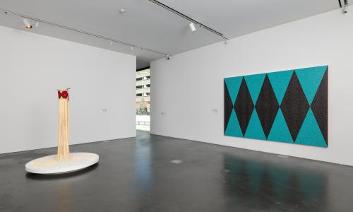 Gallery at MCA Denver with white wall and concrete-like flooring, featuring two artworks. One is large scale acrylic on canvas work featuring shades of turquoise, interstitial spaces of copper, and brownish diamond like shapes. The other is a sculpture, featuring a Copper water carrying device and ladle adorned with beadwork. Attached to the vessel are dramatic fringes/tassels.