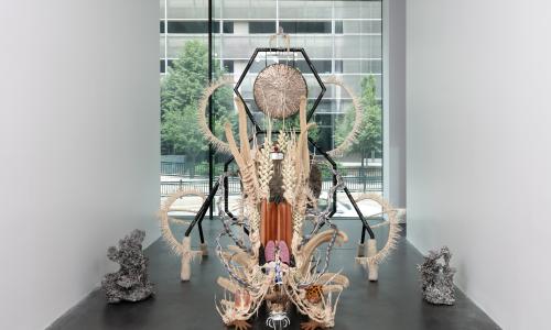 A large sculpture composed of various materials: Gong, steel, wood, cotton, glue mixture, plastic, loofah, and objects collected from a ritual of retracing the artist's original migration route. The sculpture is in a gallery space with white walls and dark flooring.