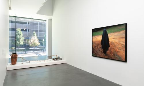 Photograph on the wall featuring a figure draped in a black sheet and standing in dirt. There is a second work in the space featuring a record player and a piece of pottery connected by what looks like a tree branch.