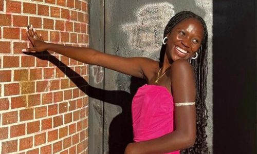 Young black female in formal attire posing with her hand on a wall, looking over her shoulder with a beaming smile. 