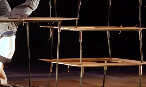 Installation of what looks like tables stacked on top of one another. A performer is doing a handstand and maneuvering their way through the installation.