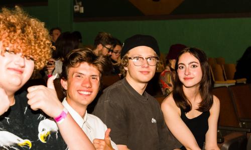 Four teens smiling and sitting in chairs in the Holiday Theater. Behind them are greenish, turquoise colored brick walls, featuring Egyptian motifs.