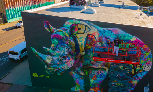 A mural by AGPNT of a multi color riNo on the side of a building with the artist, Graffiti Month, atop a left creating their mural