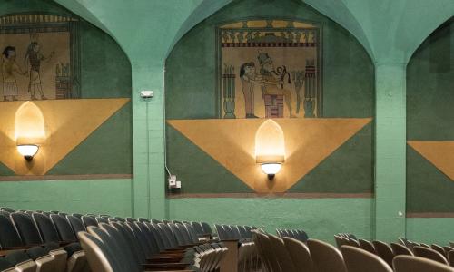 Egyptian motifs around sconces at the Holiday Theater. The walls are mostly turquoise and yellow. Rows of seats are also in view.