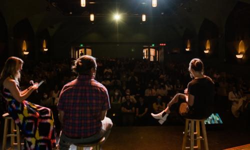 Three speakers sitting on a theater stage in front of an audience.