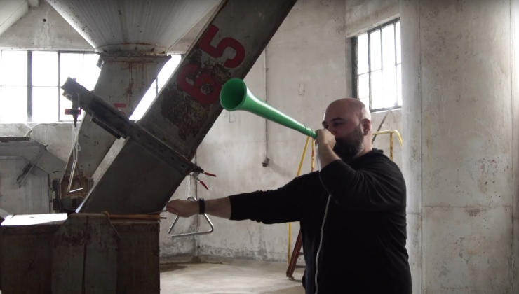 A man is blowing through a green trumpet like instrument in an indoor industrial space. 