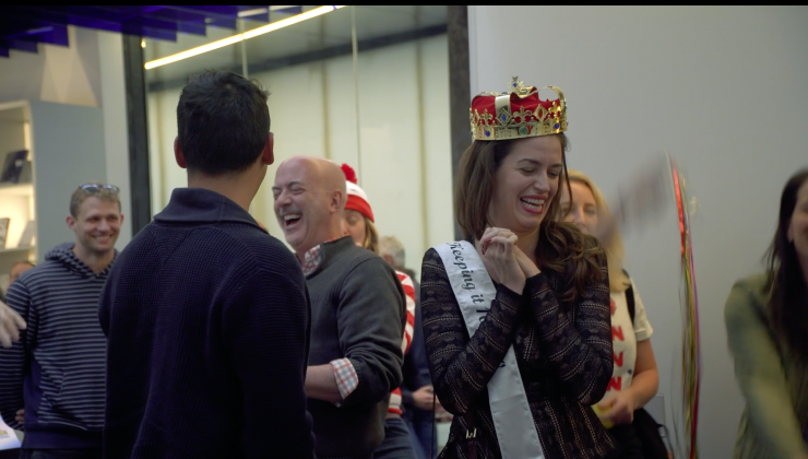 A man and a woman are surrounded by several people. The woman is wearing a white sash and a toy crown. 
