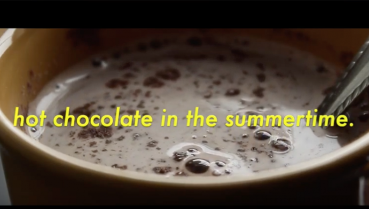 A caramel colored mug containing hot chocolate and a silver spoon with superimposed yellow text reading “hot chocolate in the summertime” 