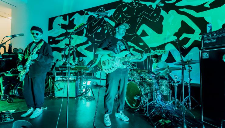 The band “Portugal the Man” performs in an MCA Denver gallery. They are lit in an aqua color and are staged in front of a large painting depicting dark abstract figures walking through a landscape of white figures bleeding.